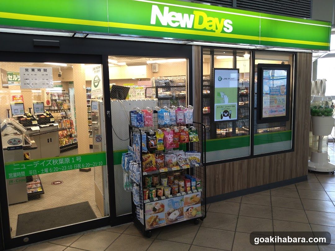 A NewDays convenience store entrance with green and white signage above the glass doors. Inside, shelves stocked with snacks and products are visible through the windows. A display rack outside holds various packaged goods.
