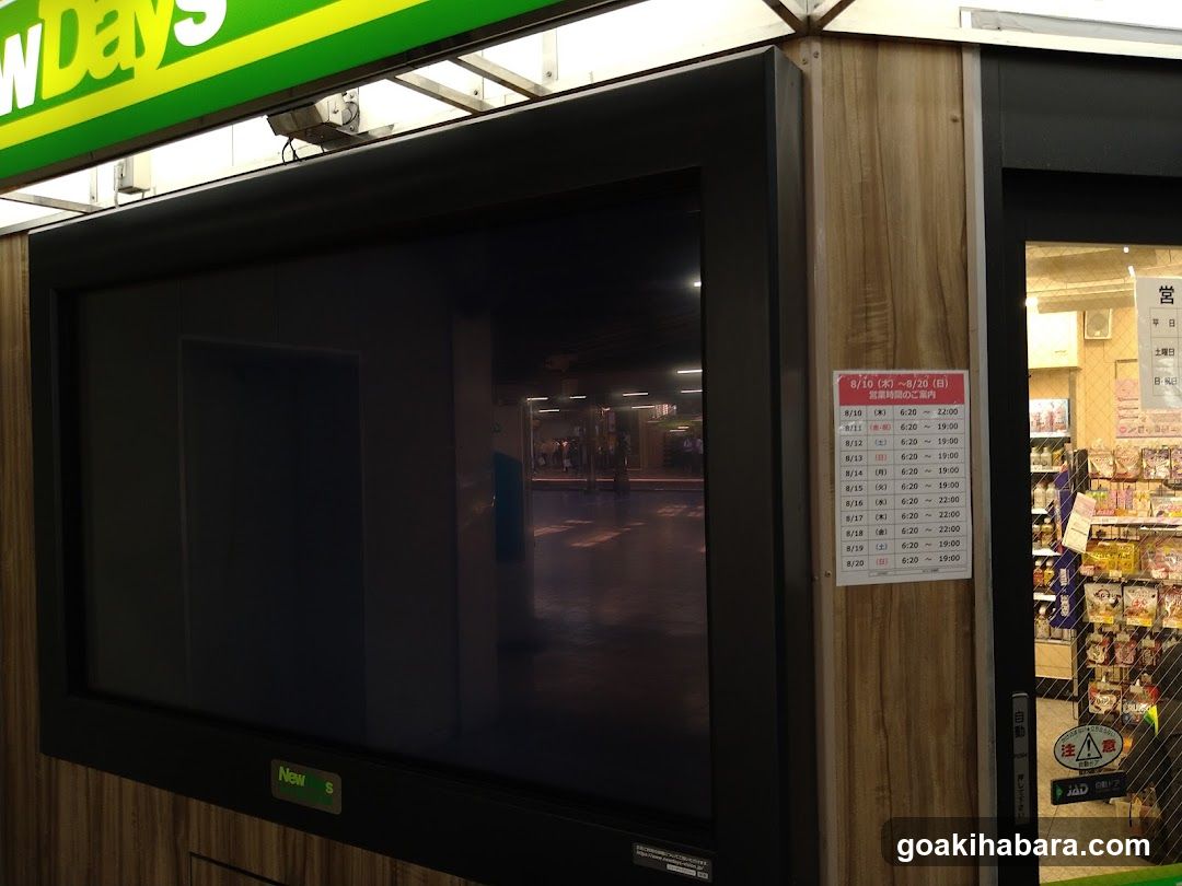 Photo of the exterior entrance showing a large digital display screen reflecting the interior, mounted on wood-paneled walls. Above is green signage reading 'NewDays'. To the right, a glass door reveals shelves stocked with packaged goods inside. A white sign lists operating hours in Japanese characters (e.g., 9:00-22:00). The scene appears to be at night.