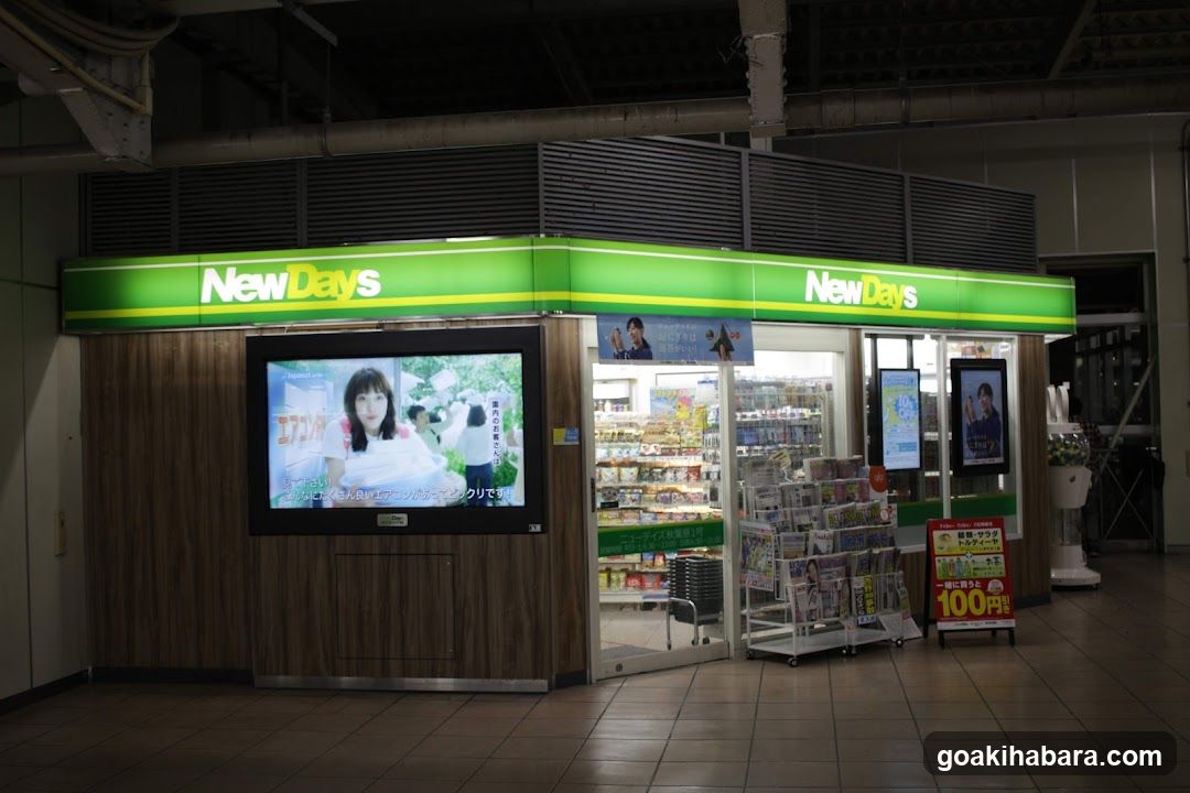 A NewDays convenience store with a green and white sign above the entrance. The storefront features a large screen displaying an advertisement, shelves stocked with products visible through glass doors, and promotional materials outside including a 100 yen discount sign.