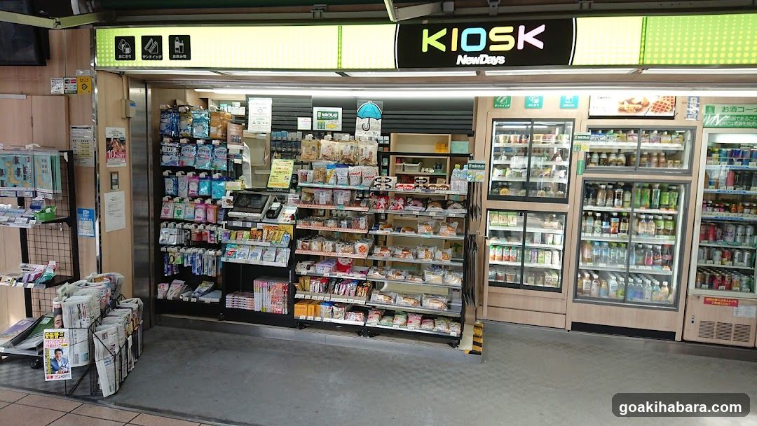 A brightly lit convenience store entrance with a 'KIOSK NowDays' sign above the door. The interior is visible, showing shelves stocked with snacks, drinks in refrigerated units, and various packaged goods. A newspaper rack stands outside near the entrance.