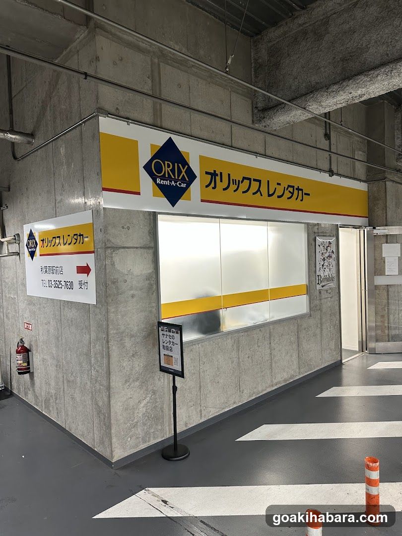 A car rental counter located in an indoor parking garage or building lobby. The storefront features a yellow and white sign with the Orix logo and Japanese text 'オリックス レンタカー'. A service window is visible, along with additional signage indicating contact information and location details.