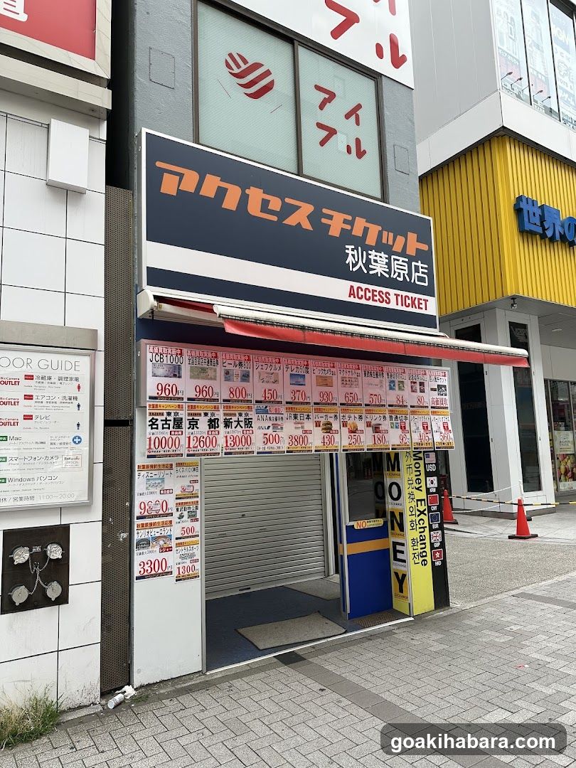 A storefront of an Access Ticket shop, featuring a large sign with Japanese and English text. The entrance is partially covered by a red awning, and various price lists are displayed on the wall.
