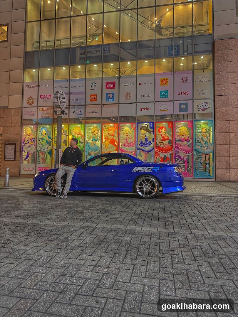 A person leaning against a modified blue sports car parked in front of a brightly lit shop facade featuring colorful anime character posters and multiple brand signs including Starbucks, ABC Mart, JINS, and others.