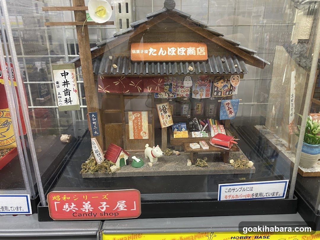 A detailed miniature model of a traditional Japanese shop with visible signage reading 'Tanbo Shop' and 'Nakai Dentaku'. The facade features a tiled roof, wooden structure, and various posters and signs attached to the walls.