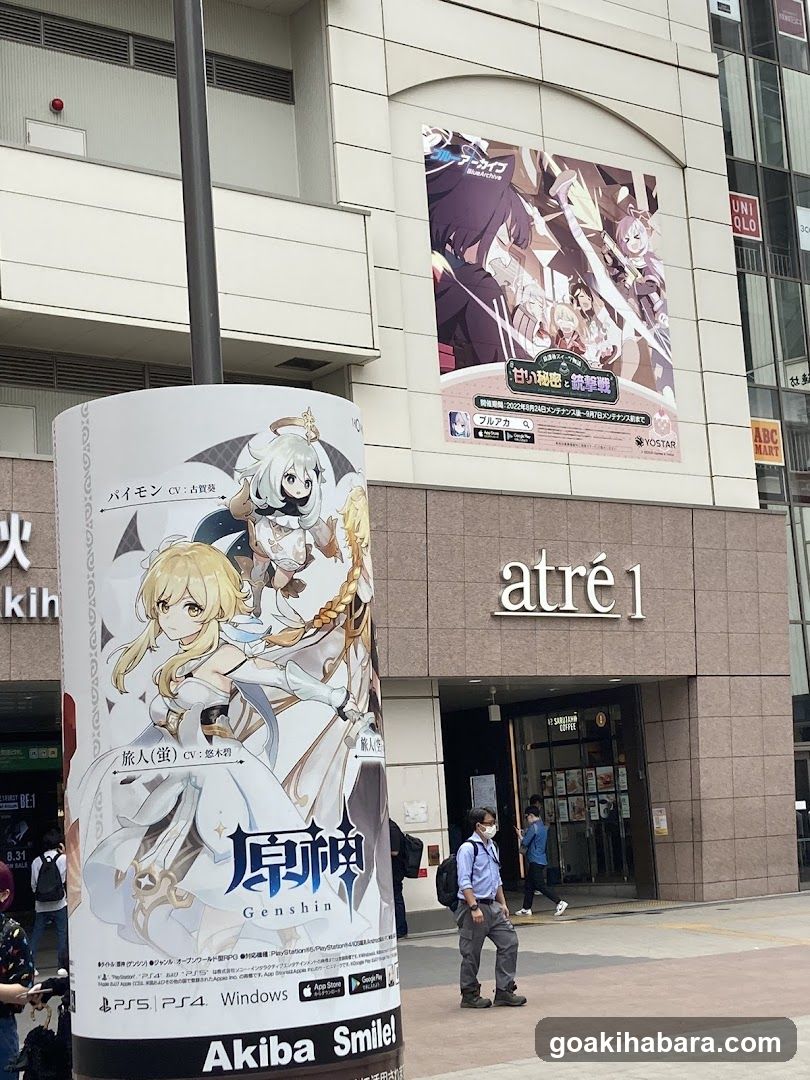 Photo of the exterior entrance to Atré1, a shopping center in Japan. The building facade features large banners advertising the game 'Genshin Impact', including characters like Baizhong and Liyue Qixing. The sign 'atré 1' is clearly visible above the glass doors where pedestrians are walking by.