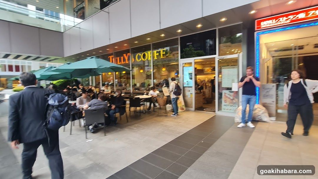 A street-level view of a Tully's Coffee storefront with outdoor seating under green umbrellas, customers dining at tables, and pedestrians walking by.
